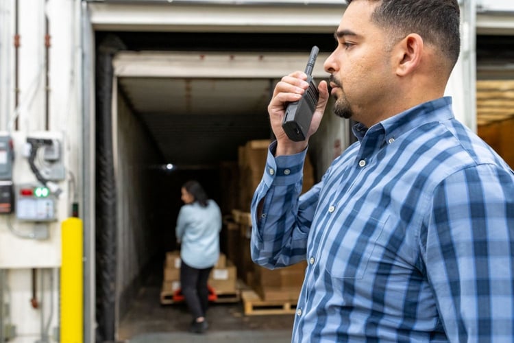 Warehouse supervisor uses a Motorola two-way radio to coordinate loading dock operations while a coworker moves shipments inside a trailer.