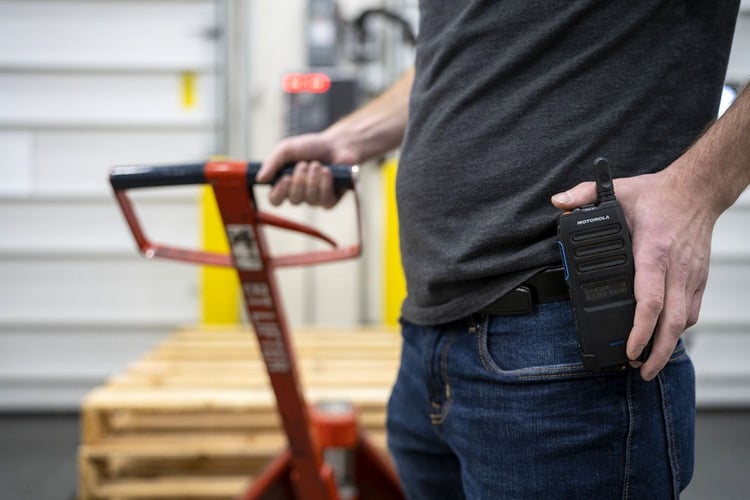 Warehouse worker with a Motorola two-way radio clipped to their belt while moving pallets with a pallet jack in a manufacturing facility.