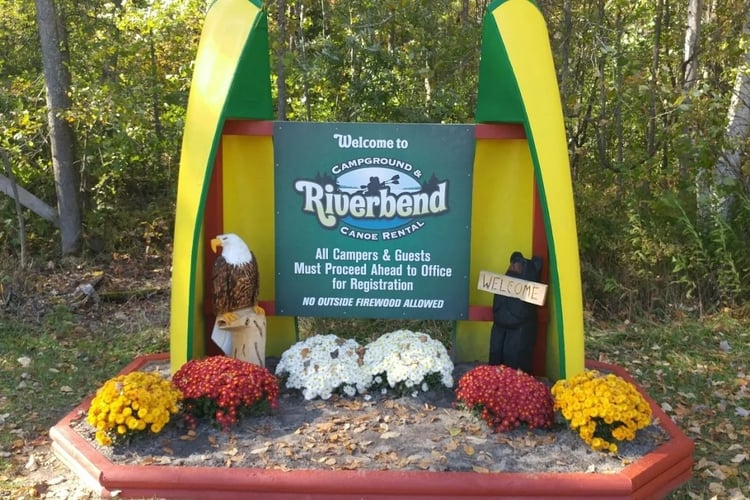Entrance sign for Riverbend Campground & Canoe Rental surrounded by trees and seasonal flowers