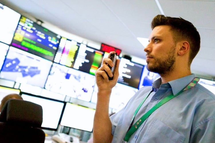 Airport operations staff member using a two-way radio in a communications control room with multiple monitoring screens displaying live operational data.