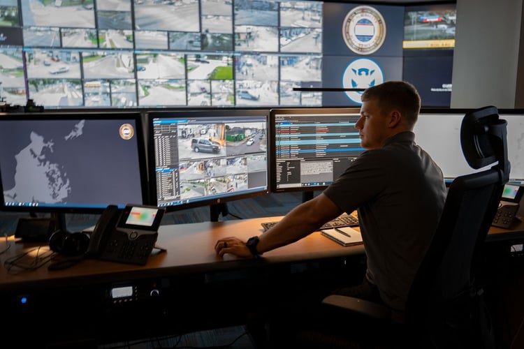 Security operations specialist monitors live camera feeds, mapping software, and dispatch systems from a multi-screen control center.