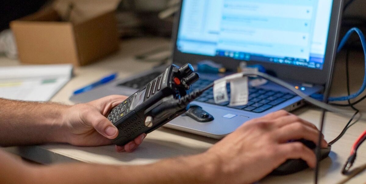 Hands holding a two-way radio connected to a laptop at a service workbench.