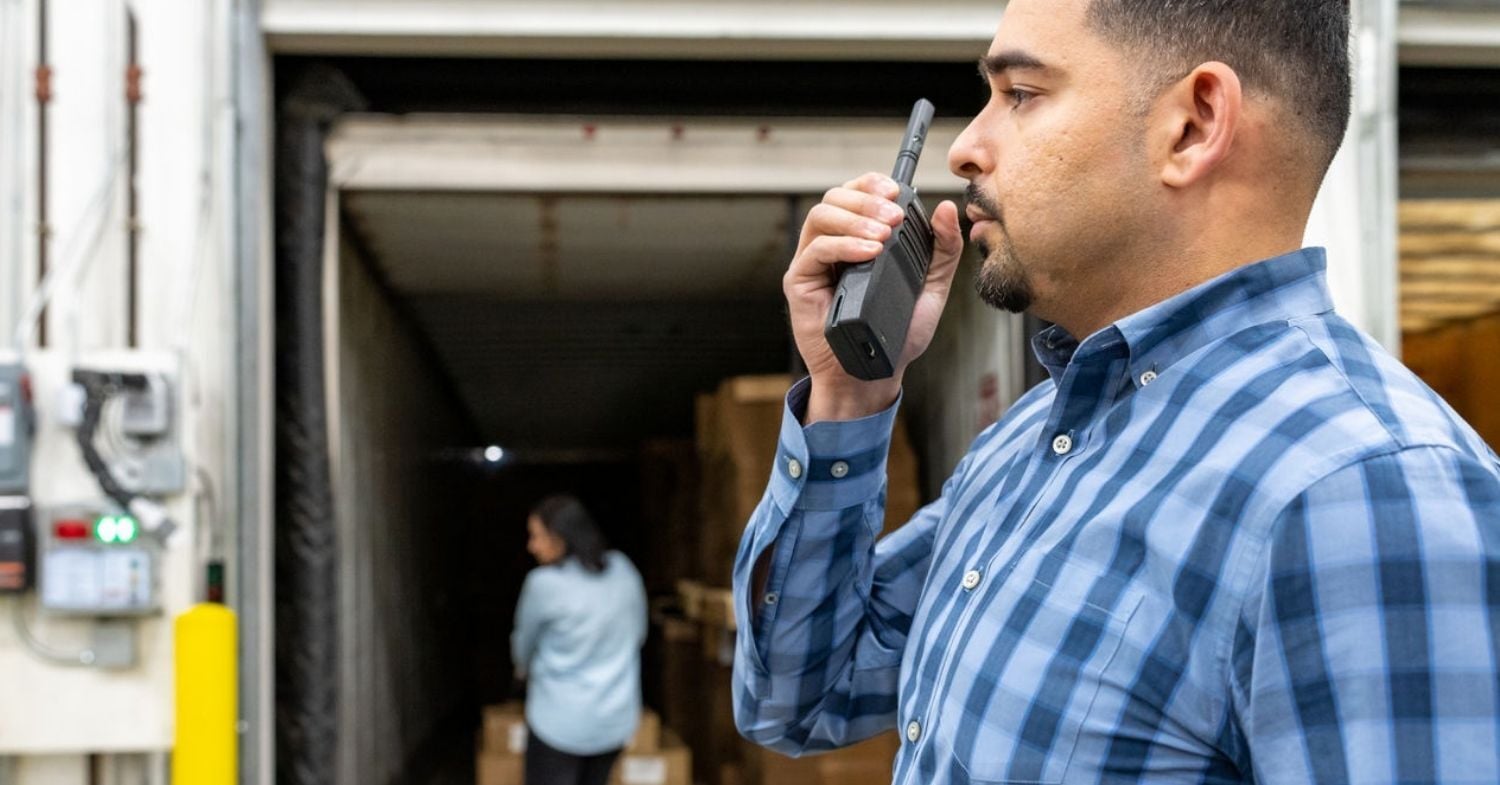 Warehouse supervisor uses a Motorola two-way radio to coordinate loading dock operations while a coworker moves shipments inside a trailer.